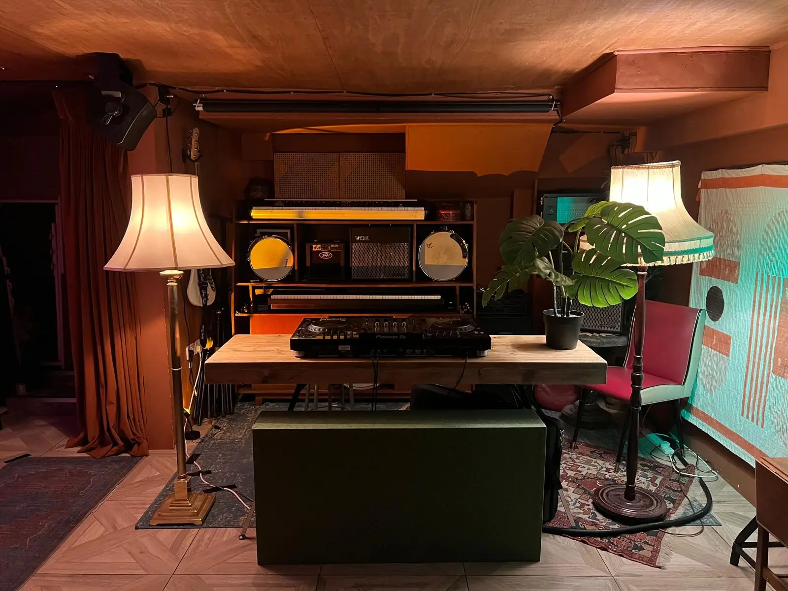 The basement, empty and lamplit: a long wooden table with DJ decks and a monstera, shelves of keyboards, drums and amps behind, a red chair and a hanging quilt