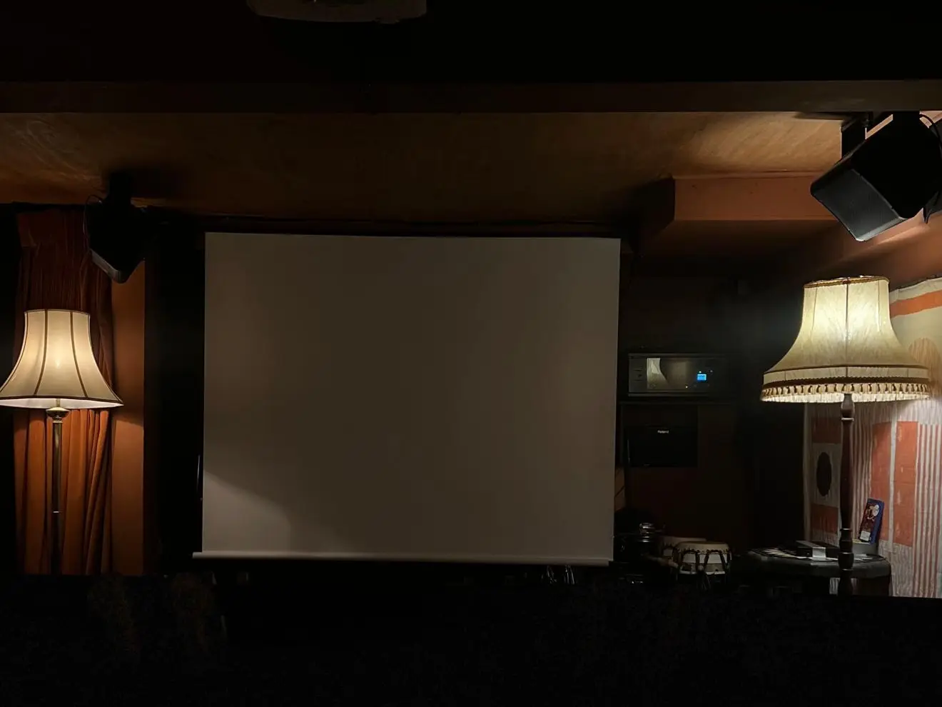 The projector screen pulled down in the basement, between two lit standard lamps