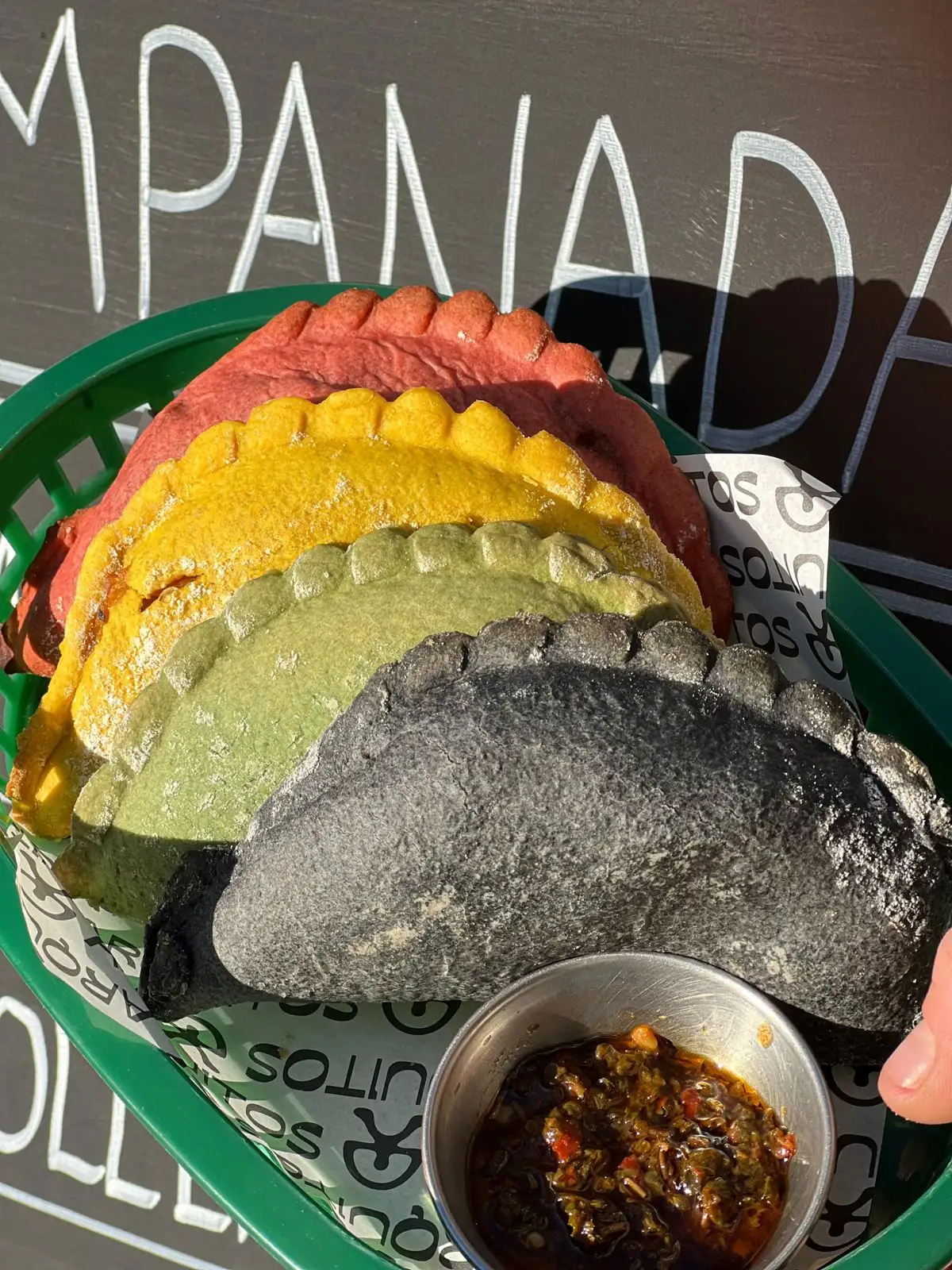 Red, yellow, green and black Arquitos empanadas in a basket, with a little pot of sauce
