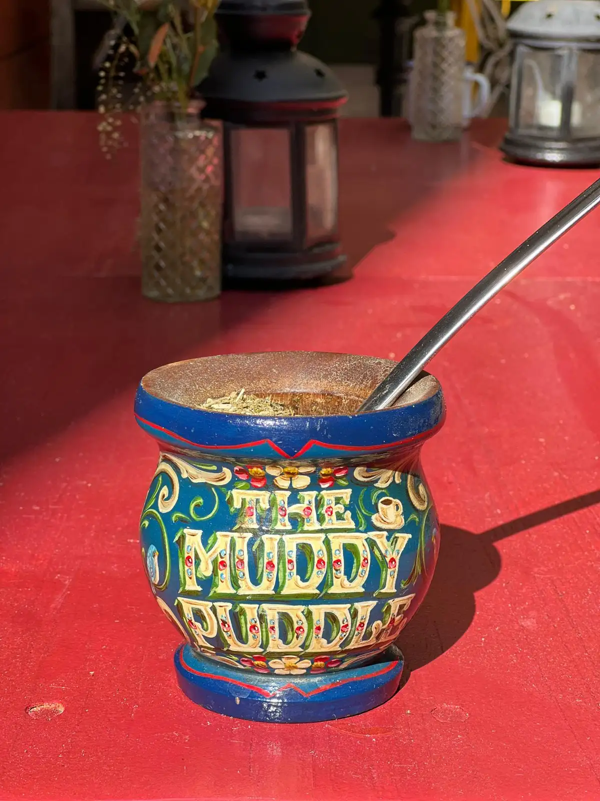 Our hand-painted Muddy Puddle mate gourd with a metal straw, on a red table in the sun