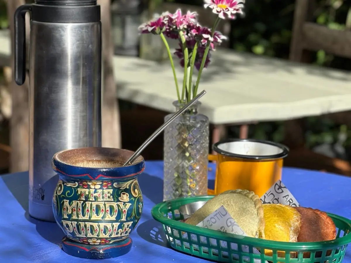 A blue table in the garden in the sun: a painted Muddy Puddle mate, a flask, flowers in a glass bottle and a basket of empanadas